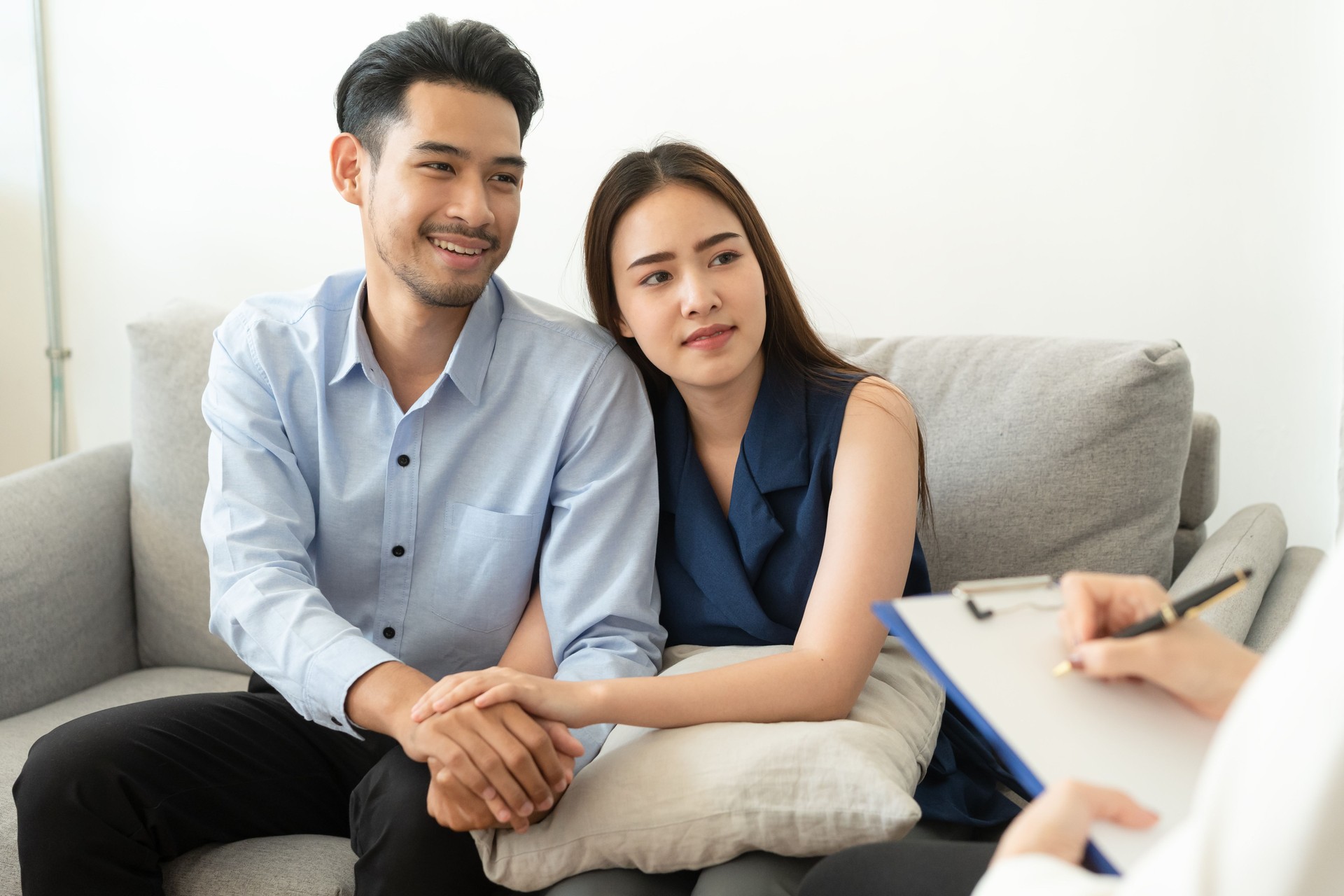 Asian couple join hand to encourage while sitting on the couch in the psychiatrist room to consult mental health problems by doctor, Health and illness concepts